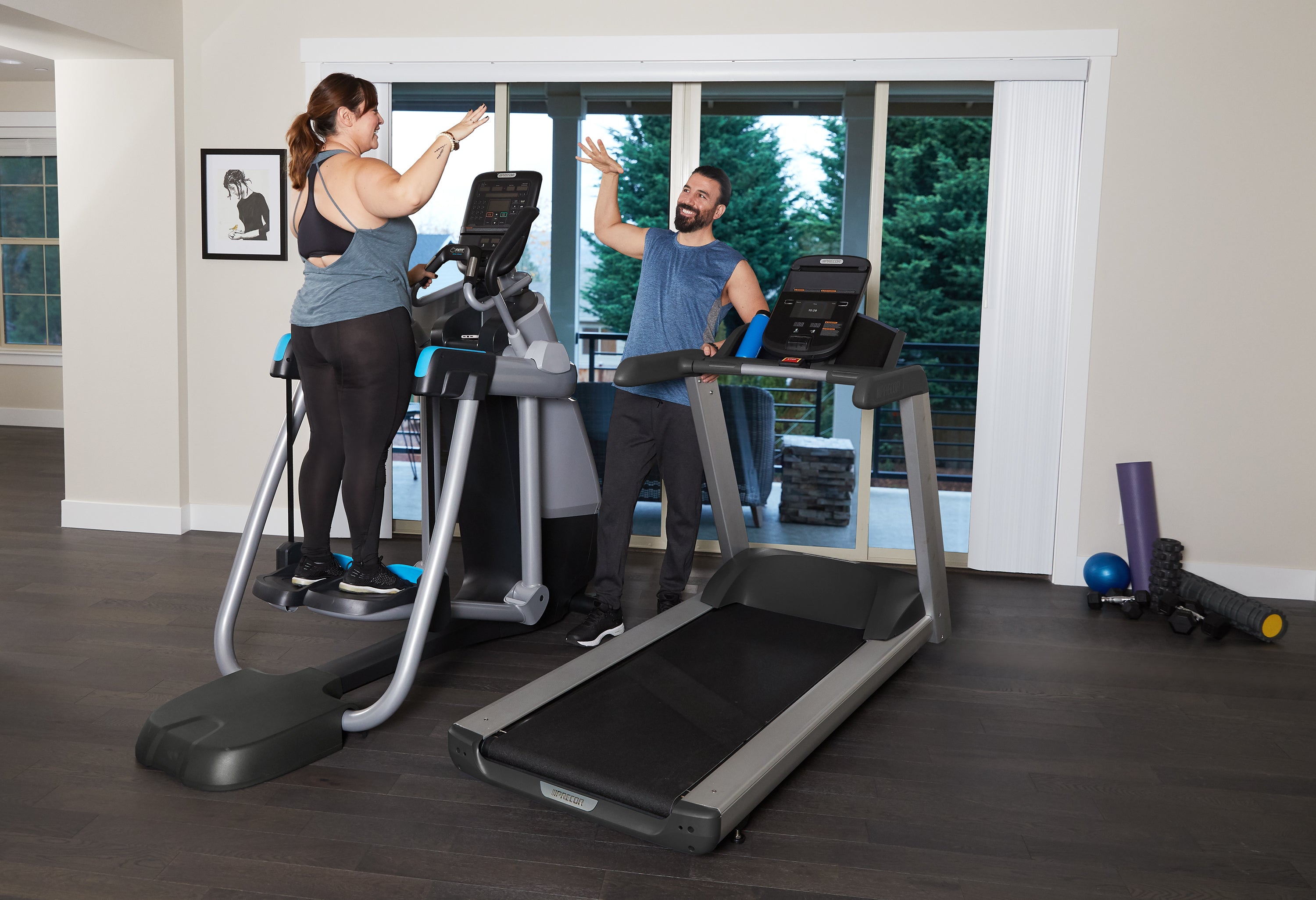 Couple high fiving each other after a workout in their home gym. Female is on a Precor AMT 835 Adaptive Motion Trainer with P31 console and the man is leaning on the TRM 445 treadmill