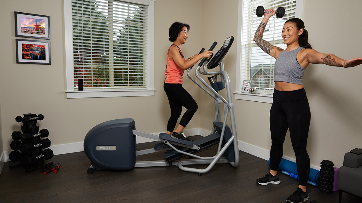Two women working out at home, one is lifting a dumbbell over her head, the other is on a Precor elliptical 