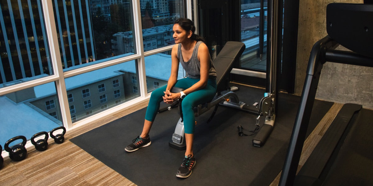 Female resting between sets in her home gym that features Precor DBR 119 adjustable bench, FTS Glide functional strength training system, and a Precor TRM treadmill