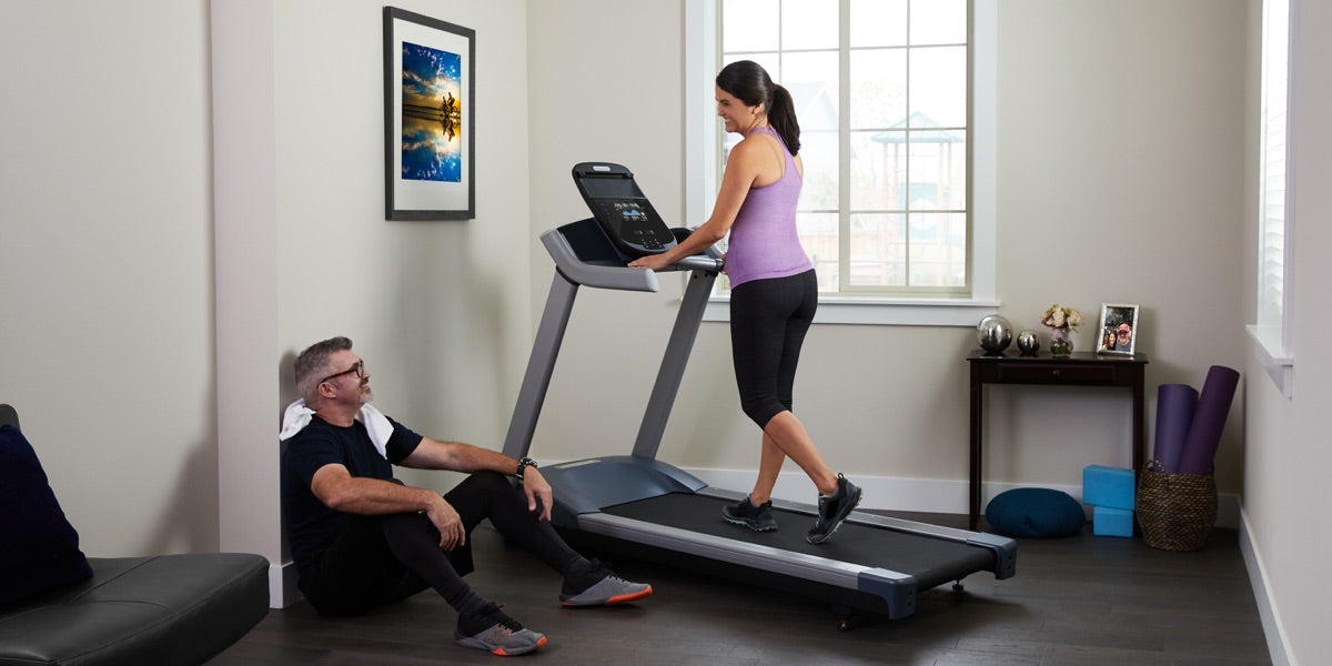 Woman walking on a Precor treadmill at home with her husband chilling on the floor post-workout