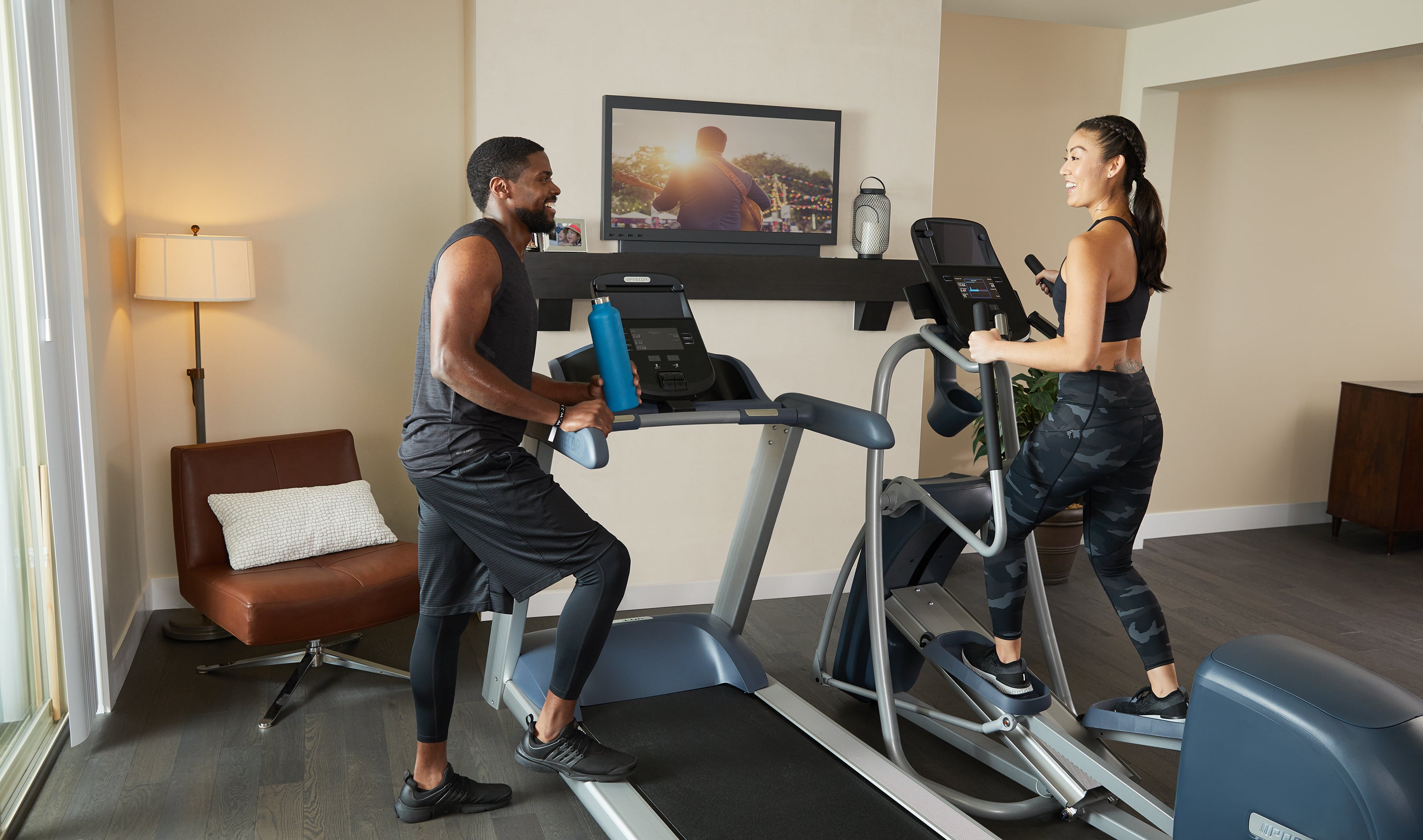Woman working out on Precor EFX 447 elliptical next to a Precor TRM 445 treadmill with man leaning on arm of treadmill chatting after workout at home