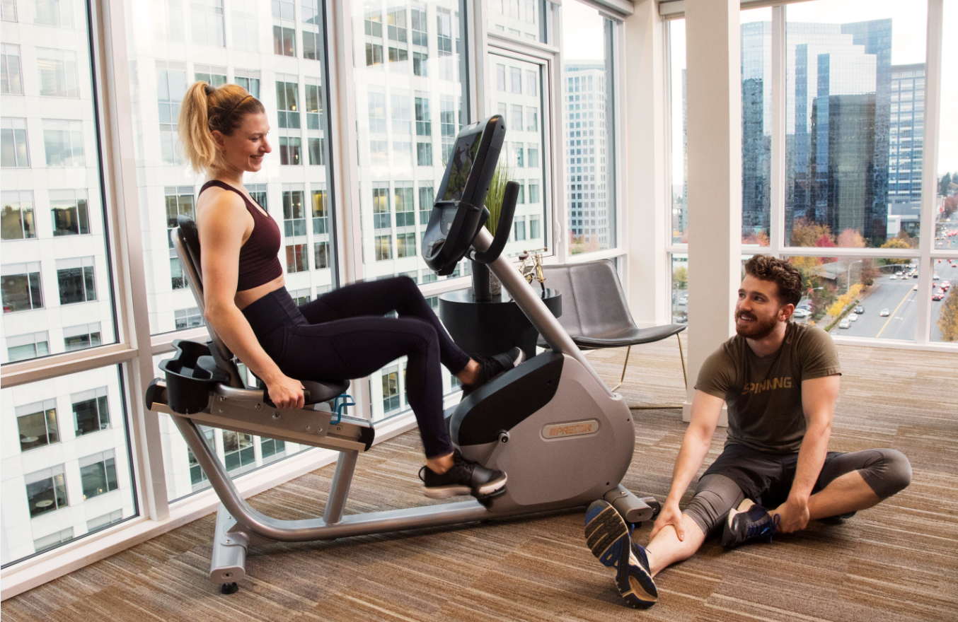 Female on a Precor RBK 865 recumbent bike with a man on the floor stretching